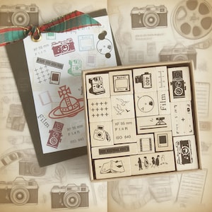 May include: A collection of wooden stamps featuring camera and film-related designs. The stamps are in a box, alongside a tag with similar illustrations and a red and green ribbon. The text includes 'Film'.