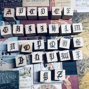 Puede incluir: Un conjunto de veintiséis bloques de madera cuadrados, cada uno con una letra negra del alfabeto impresa en una tipografía gótica decorativa. Los bloques están dispuestos en filas, mostrando la secuencia completa de la A a la Z de forma uniforme.