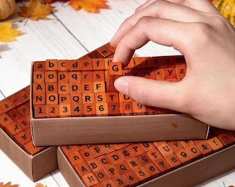 Puede incluir: Sellos de madera con letras y números en una caja marrón. Los sellos están dispuestos en una cuadrícula, con letras y números visibles. Una mano sostiene un sello con la letra "G". El fondo incluye hojas de otoño y calabazas.