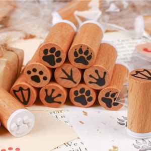 May include: A selection of wooden stamps featuring animal paw and bird foot prints. The stamps have a natural wood grain finish and black ink imprints. The stamps are displayed on a light-toned surface, suitable for crafting or stationery projects.