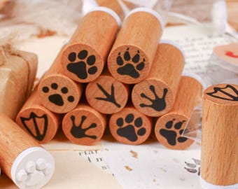 May include: A collection of wooden stamps with various animal paw and bird foot designs. The stamps have a natural wood finish and black ink imprints. The stamps are arranged on a light-colored surface, suggesting a craft or stationery theme.