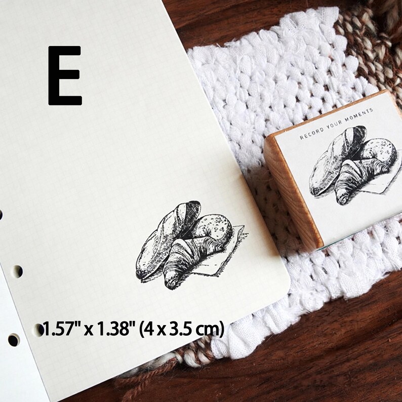 May include: A wooden rubber stamp featuring a black ink illustration of a baguette, a croissant, and a round bread roll. The stamp includes the text "RECORD YOUR MOMENTS" and measures 1.57 inches by 1.38 inches. The stamp is shown with a sample print.