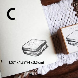 May include: A wooden rubber stamp featuring a black ink illustration of a sandwich, with the text 'RECORD YOUR M' visible on the side. The stamp impression on the paper measures 1.57 inches by 1.38 inches, labeled with the letter C.