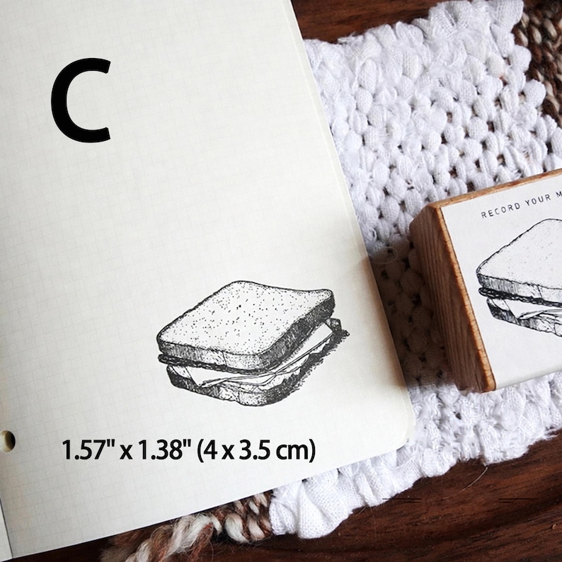 May include: A wooden rubber stamp featuring a black ink illustration of a sandwich, with the text 'RECORD YOUR M' visible on the side. The stamp impression on the paper measures 1.57 inches by 1.38 inches, labeled with the letter C.