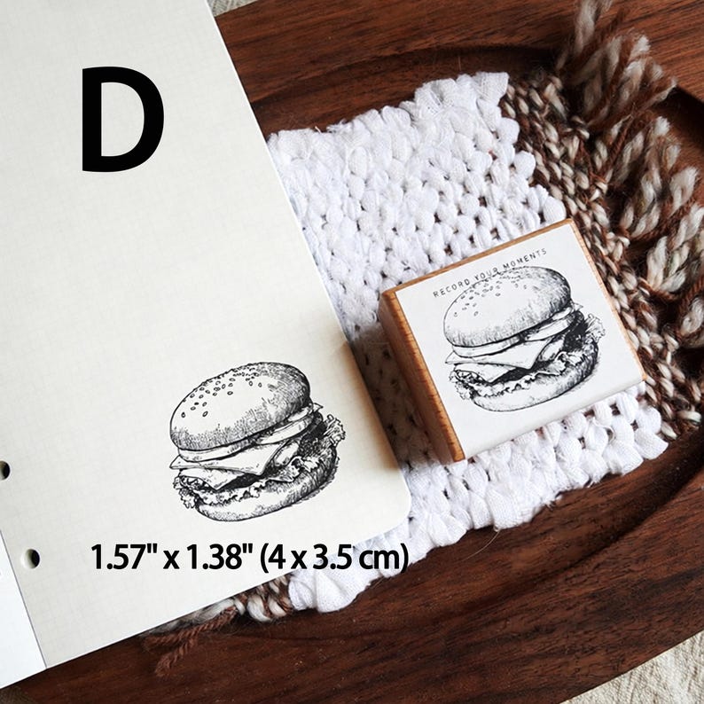 May include: A wooden rubber stamp featuring a detailed black and white line art illustration of a hamburger with the text "RECORD YOUR MOMENTS" printed above it. The stamp measures 1.57 inches by 1.38 inches and is shown with a stamped impression.