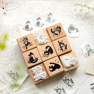 Könnte beinhalten: Eine Sammlung von Holzstempeln mit Illustrationen von schwarzen Katzen. Die Stempel zeigen Katzen in verschiedenen Posen, darunter Katzen mit Hexenhüten und beim Lesen von Büchern. Einige Stempel sind auf Papier abgedruckt.