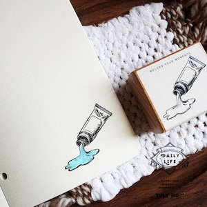 May include: A wooden rubber stamp featuring a black and white illustration of a paint tube with the text RECORD YOUR MOMENTS. The stamp is shown next to a grid paper page with a stamped image of the paint tube and a blue puddle of paint.