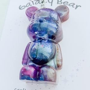May include: A resin bear-shaped keyring with a galaxy design in purple, blue, and pink hues. The keyring has a silver metal loop at the top. The text "Galaxy Bear" is visible.