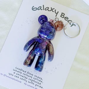 以下が含まれることがあります： ギャラクシーベアのキーホルダー。クマの形をしたチャームは、紫、青、金色の樹脂の混合物で、キーリングが付いています。「Galaxy Bear」という文字がカードの上部にあります。カードには「混沌から作られ、色が衝突し、宇宙が捉えられ、誇りを持って持ち運ぶことができます」というテキストも含まれています。