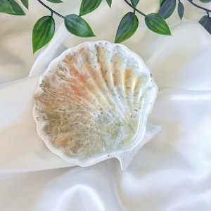 May include: A seashell-shaped decorative tray with a glossy finish. The tray features a blend of white, orange, and green hues, resembling a natural seashell. The tray is placed on a white, silky fabric, with green leaves in the background.