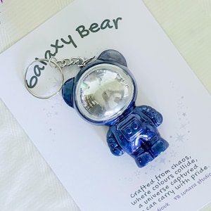 Könnte beinhalten: Ein blauer Astronauten-Bären-Schlüsselanhänger mit silbernem Visier. Der Schlüsselanhänger befindet sich auf einer weißen Karte mit dem Text "Galaxy Bear" und einem Zitat über das Tragen eines Universums mit Stolz.