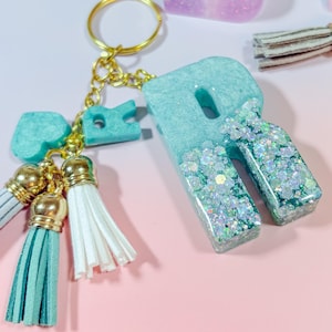 May include: A teal resin letter "R" keychain with gold hardware and a gold keyring. The letter is filled with glitter and has a matching heart-shaped charm and tassels in white and teal. The background is pink.
