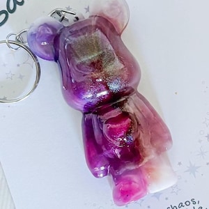 May include: A purple and pink resin bear keyring with a silver ring. The bear-shaped charm has a glittery, iridescent effect. The keyring is a decorative accessory.