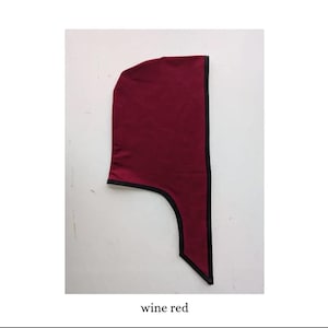May include: A wine red hood featuring a structured, pointed top and a long, tapered neck piece. The edges of the hood are finished with a thin, contrasting black trim that outlines the entire shape of the garment against the plain background.