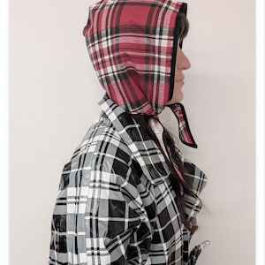 May include: A headscarf featuring a red, black, and white plaid pattern with a black trim, worn with a matching trench coat that displays a black, grey, and white plaid pattern. The coat has a glossy finish, a wide collar, and a belted waist.
