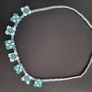 Puede incluir: Un collar de cuentas con diseños florales turquesa y blancos. El collar está ensartado en una cadena de color plateado y tiene un cierre de broche. Las flores están espaciadas uniformemente a lo largo del collar.