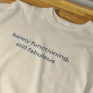 Puede incluir: Una camiseta color crema con el texto "barely functioning, still fabulous" impreso en azul oscuro. La camiseta está colocada sobre una superficie de madera, mostrando su diseño y color.