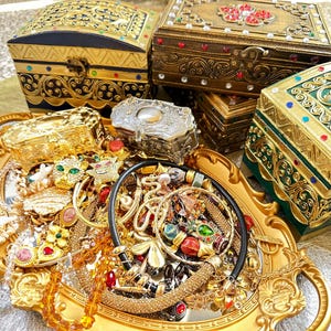 May include: A collection of decorative jewellery boxes and a gold-coloured tray filled with necklaces, bracelets, and rings. The boxes are in gold, green, and black, and are adorned with jewels.