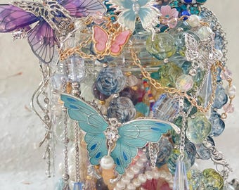 Peut inclure: Un bocal en verre transparent rempli d'un assortiment de perles colorées et de papillons décoratifs. Les papillons sont dans des tons de violet, rose et bleu, avec des détails en métal. Le bocal déborde de bijoux et d'éléments décoratifs.