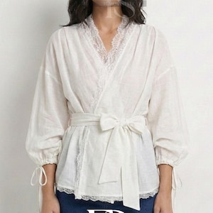 Op de afbeelding: Een witte wikkelblouse met een delicate kanten rand langs de halslijn en zoom. De blouse heeft lange mouwen met strikdetails bij de manchetten en een bijpassende stoffen ceintuur die in de taille wordt gestrikt. De blouse wordt gecombineerd met een blauwe jeans.