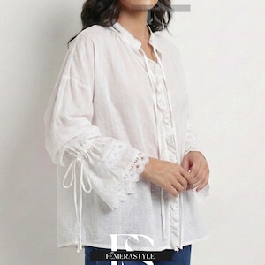 Op de afbeelding: Een witte blouse met lange mouwen en kanten details op de manchetten. De blouse heeft een opstaande kraag en een striksluiting bij de hals. De mouwen zijn bij de polsen met strikjes gerimpeld. De blouse wordt gecombineerd met een blauwe jeans.