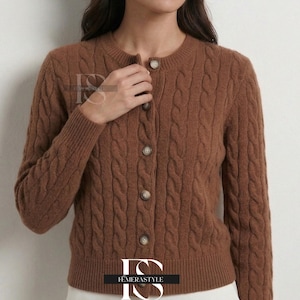 Op de afbeelding: Bruine kabelgebreide cardigan met een ronde hals en knoopsluiting. De lange mouwen trui heeft een klassiek design en is gemaakt van een zacht materiaal. De cardigan is een veelzijdig kledingstuk om in laagjes te dragen.