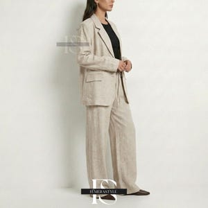 May include: A beige linen blazer and matching wide-leg trousers set. The blazer features a notched lapel, long sleeves, and two front flap pockets. The trousers have a relaxed, straight-leg fit. A black top is worn underneath the blazer.
