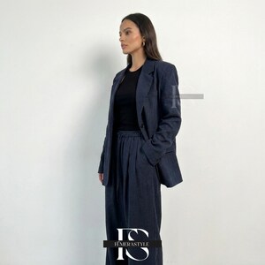 May include: A dark charcoal gray blazer and matching wide-leg trousers with a drawstring waist, worn over a plain black crew-neck top. The blazer features notched lapels, a single button closure, and long sleeves with buttons at the cuffs.