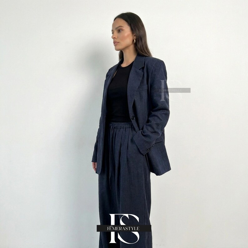 May include: A dark charcoal gray blazer and matching wide-leg trousers with a drawstring waist, worn over a plain black crew-neck top. The blazer features notched lapels, a single button closure, and long sleeves with buttons at the cuffs.