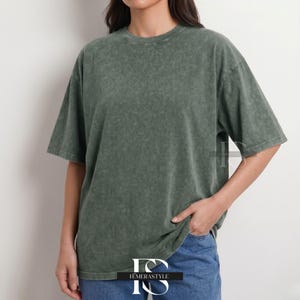 Pu&ograve; includere: Una maglietta verde oliva sbiadita con girocollo e maniche corte. La maglietta ha una vestibilit&agrave; leggermente oversize e un aspetto strutturato. La modella indossa jeans blu. Il logo "IEMERASTYLE" &egrave; visibile in basso.