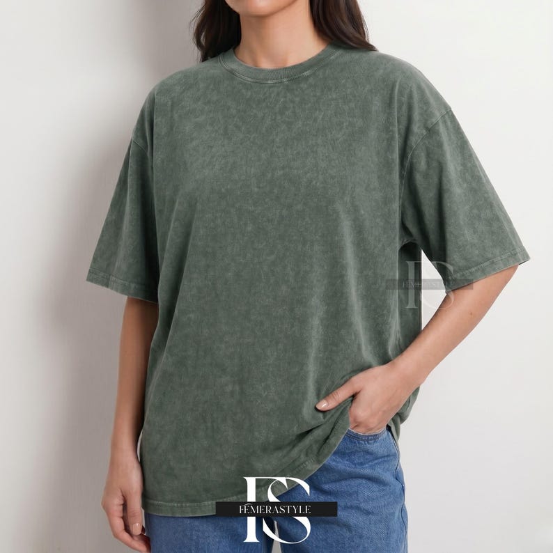 Pu&ograve; includere: Una maglietta verde oliva sbiadita con girocollo e maniche corte. La maglietta ha una vestibilit&agrave; leggermente oversize e un aspetto strutturato. La modella indossa jeans blu. Il logo "IEMERASTYLE" &egrave; visibile in basso.