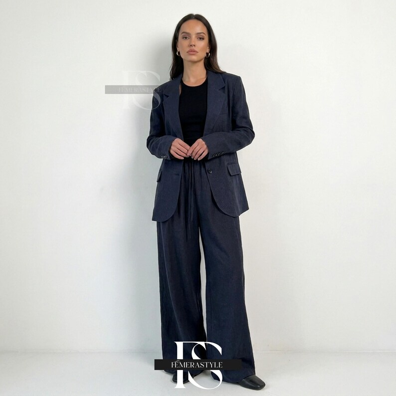 May include: A dark navy blue blazer and matching wide-leg trousers set worn over a black sleeveless top. The blazer features a notched lapel, two front buttons, and two flap pockets. The trousers have a high-waisted fit with a drawstring waist.