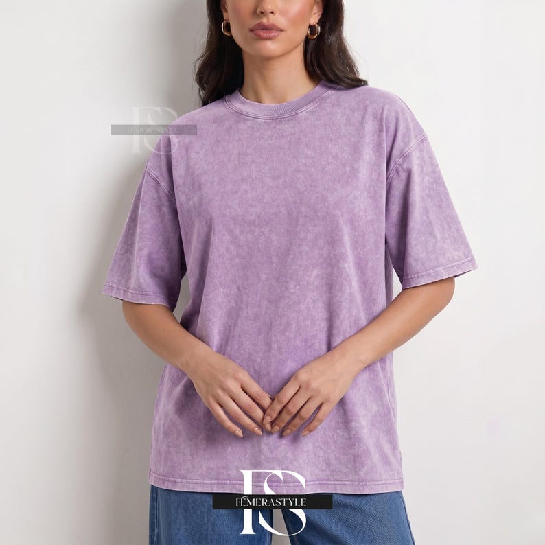 Pu&ograve; includere: Una maglietta lilla a maniche corte con scollo tondo. La maglietta ha un aspetto leggermente sbiadito e strutturato. La modella indossa jeans blu. Il nome del marchio "IEMERASTYLE" &egrave; visibile in basso nell'immagine.