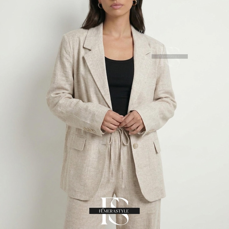May include: A beige blazer with a textured finish, featuring notched lapels, long sleeves, and a single button closure. The blazer has two front flap pockets and is worn over a black tank top, paired with matching beige trousers with a drawstring waist.