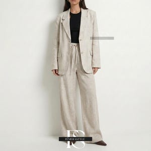 May include: A beige blazer and matching wide-leg trousers set featuring a textured fabric. The blazer has notched lapels, two front buttons, and flap pockets. The trousers include a drawstring waist. A black sleeveless top is worn underneath.