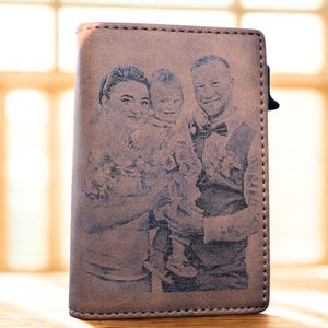 May include: A brown leather wallet with a family portrait engraved on the front. The portrait depicts a woman, a man wearing a bow tie, and a child. The wallet has a stitched border and a black clasp.
