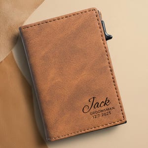 May include: A brown leather wallet with the name "Jack" and the text "Groomsman 12.7.2025" printed on the front. The wallet has a stitched border and a black clasp. The background is a light brown color.