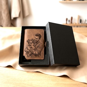 May include: A tan leather wallet featuring a laser-engraved portrait of a person holding a dog, presented inside a black rectangular gift box with a matching black lid, resting on a light-colored fabric surface.