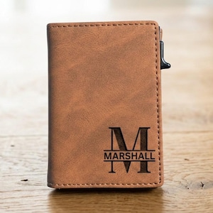 May include: A brown leather wallet with the name "Marshall" and the letter "M" engraved on the front. The wallet has a stitched border and a black metal clip on the side. The wallet is sitting on a wooden surface.