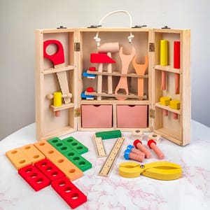 Op de afbeelding: Een houten speelgoedgereedschapskist met een wit touw handvat, open om speelgoedgereedschap in verschillende kleuren te onthullen. Inclusief een rode zaag, hamer, moersleutel en andere gereedschappen. Extra speelgoedblokken, linialen en schroeven zijn vooraan gerangschikt.