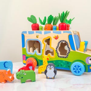 Op de afbeelding: Een houten speelgoedbus met kleurrijke dierenblokken en wortelvormige toppers. De bus heeft uitsparingen voor de blokken, waaronder een giraf, een olifant en een pinguïn. De bus is beschilderd met groene, blauwe en gele accenten.