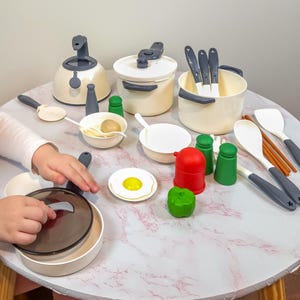 Op de afbeelding: Een speelkeuken set met diverse speelgoed kookgerei en voedsel items. De set bevat een waterkoker, potten, pannen, lepels en speelgoed voedsel zoals een ei en peper. Het speelgoed is voornamelijk crème en grijs, met rode en groene accenten.
