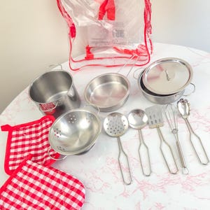 May include: A toy kitchen set made of stainless steel, featuring pots, pans, and utensils, alongside red and white gingham oven mitts and pot holders. The set is packaged in a clear plastic bag with red edging.