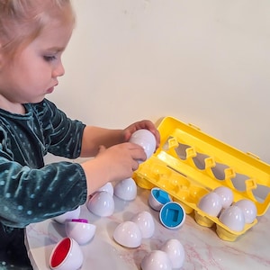 Può includere: Un bambino gioca con un set di uova di plastica bianche e un cartone per uova giallo. Alcune uova sono aperte, rivelando interni colorati rossi, blu e viola. Le uova sono su un tavolo con motivo marmorizzato bianco e rosa.