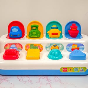 May include: A colorful pop-up toy featuring various vehicles, including a car, train, and boat. The toy has a white base with blue accents and includes red, yellow, green, and blue components. The toy is designed for young children.