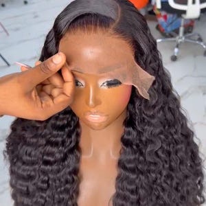 May include: A dark-haired wig with long, curly locks is displayed on a mannequin. The wig features a lace front and is being styled. The mannequin has a dark complexion and is wearing makeup. The background shows a salon setting.