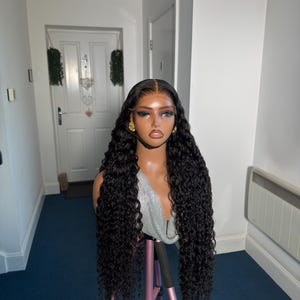 May include: A black curly wig on a mannequin head. The wig is long and has a natural-looking hairline. The mannequin is on a pink tripod stand. The background is a hallway with a white door and blue carpet.