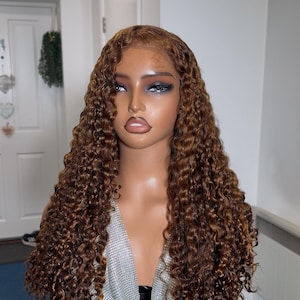 May include: A mannequin head showcasing a long, curly wig. The wig is a warm brown shade with lighter strands, and the curls are tightly wound. The mannequin is wearing a silver, sparkly top. The background is a room with a white door and walls.