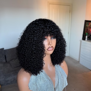 May include: A black curly wig with a fringe, styled on a mannequin head. The mannequin is wearing a silver, sparkly halter top. The background is a room with a couch and a door.