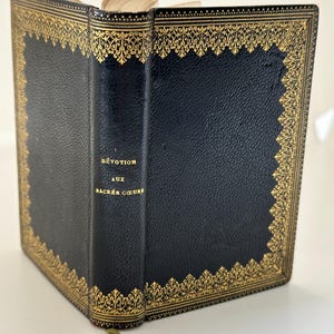 May include: A vintage book with a dark blue cover and gold decorative trim. The book's spine features the title "Dévotion aux Sacrés Coeurs" in gold lettering. A gold-colored ribbon bookmark is visible.